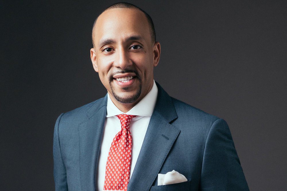 Headshot on a dark grey background of a sminling professional black man wearing a blue suit with red tie, a well groomed goatee and short cut hair.
